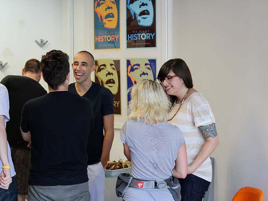 A group of people are standing and conversing in a room. A man and a woman are smiling and laughing near the center. In the background, there are framed posters on the wall featuring a stylized portrait of a person and the text "We Made History.
