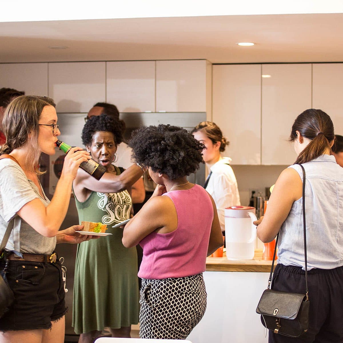 People socializing at an indoor gathering. A woman on the left is eating while holding a drink and talking to another individual. The setting appears to be a casual, community or office event, with conversations happening around a kitchen counter with refreshments.