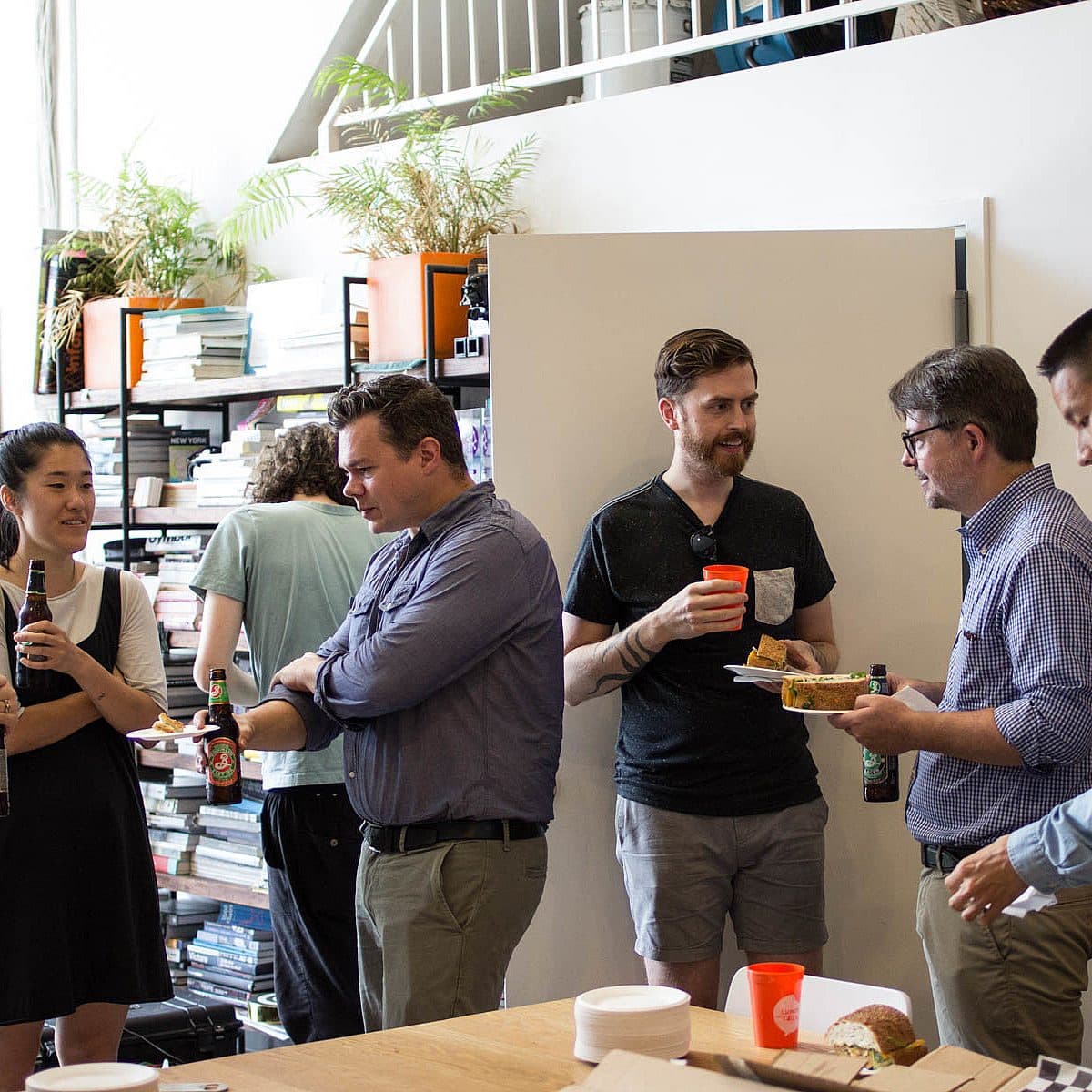 People socializing at a casual indoor gathering. Some are holding drinks and food, while others engage in conversation. There's a mix of men and women, with books and plants visible in the background. A table with more food and drinks is in the foreground.