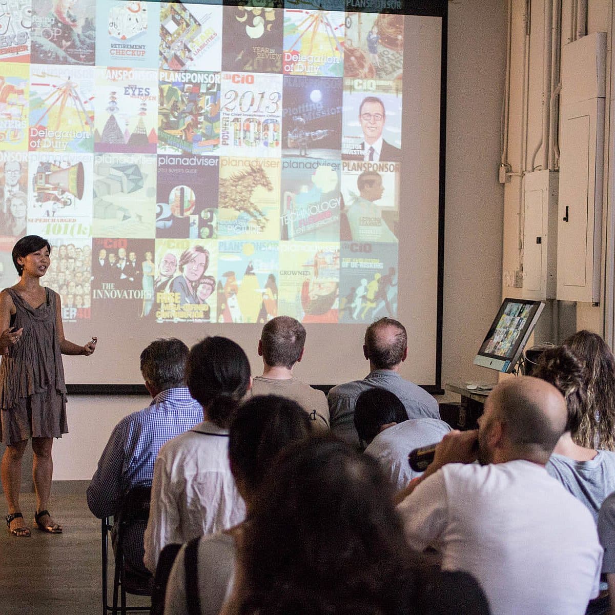 A woman stands and presents to an audience in a room, with a large screen behind her displaying a collage of posters and magazine covers. The audience is seated and attentively listening. The room has a casual setting with white walls and various decorations.