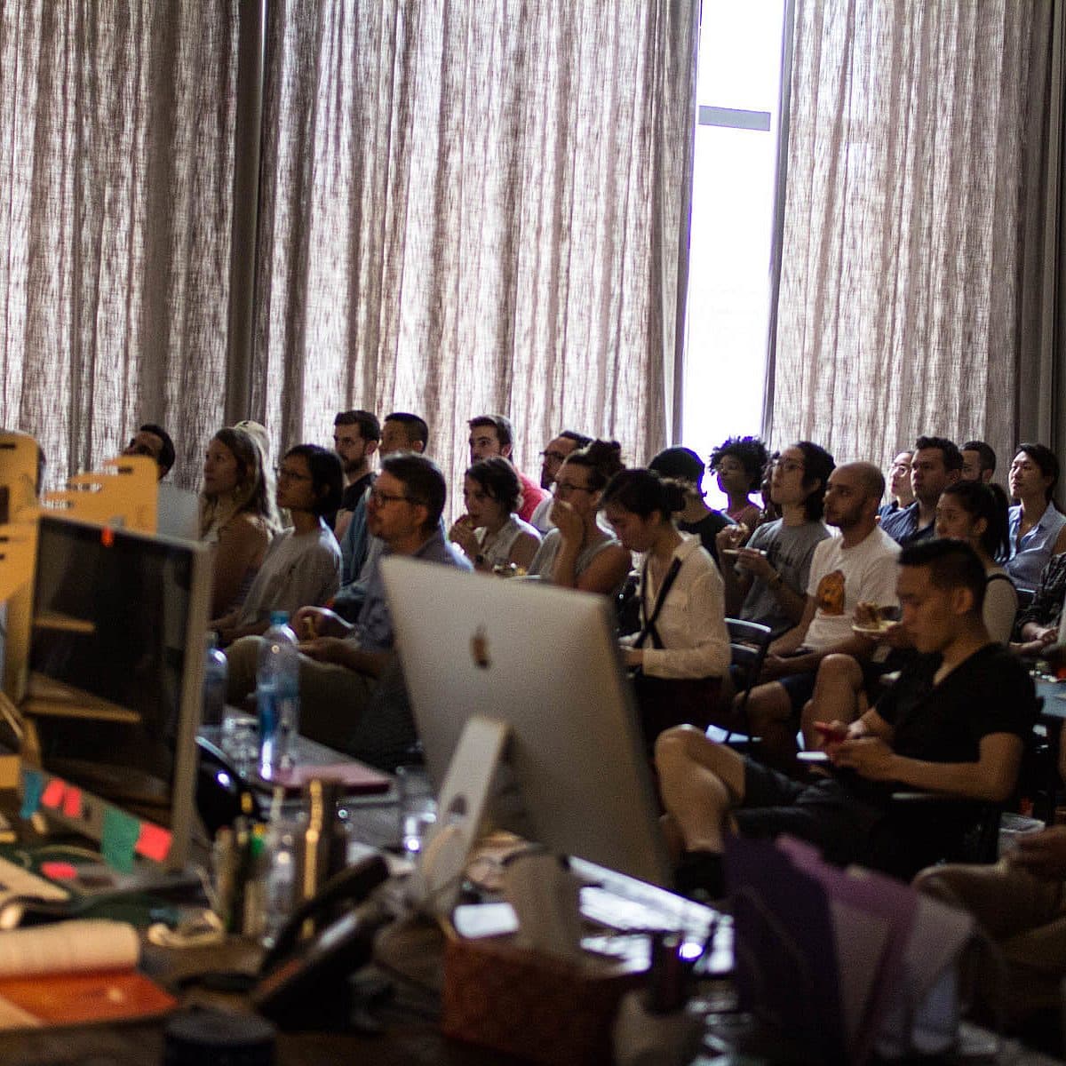 A diverse group of individuals are attentively listening to a presentation in a large room. Several computer monitors and equipment are visible in the foreground, while the audience is seated in rows, facing towards the unseen speaker. The room has large windows with drawn curtains.