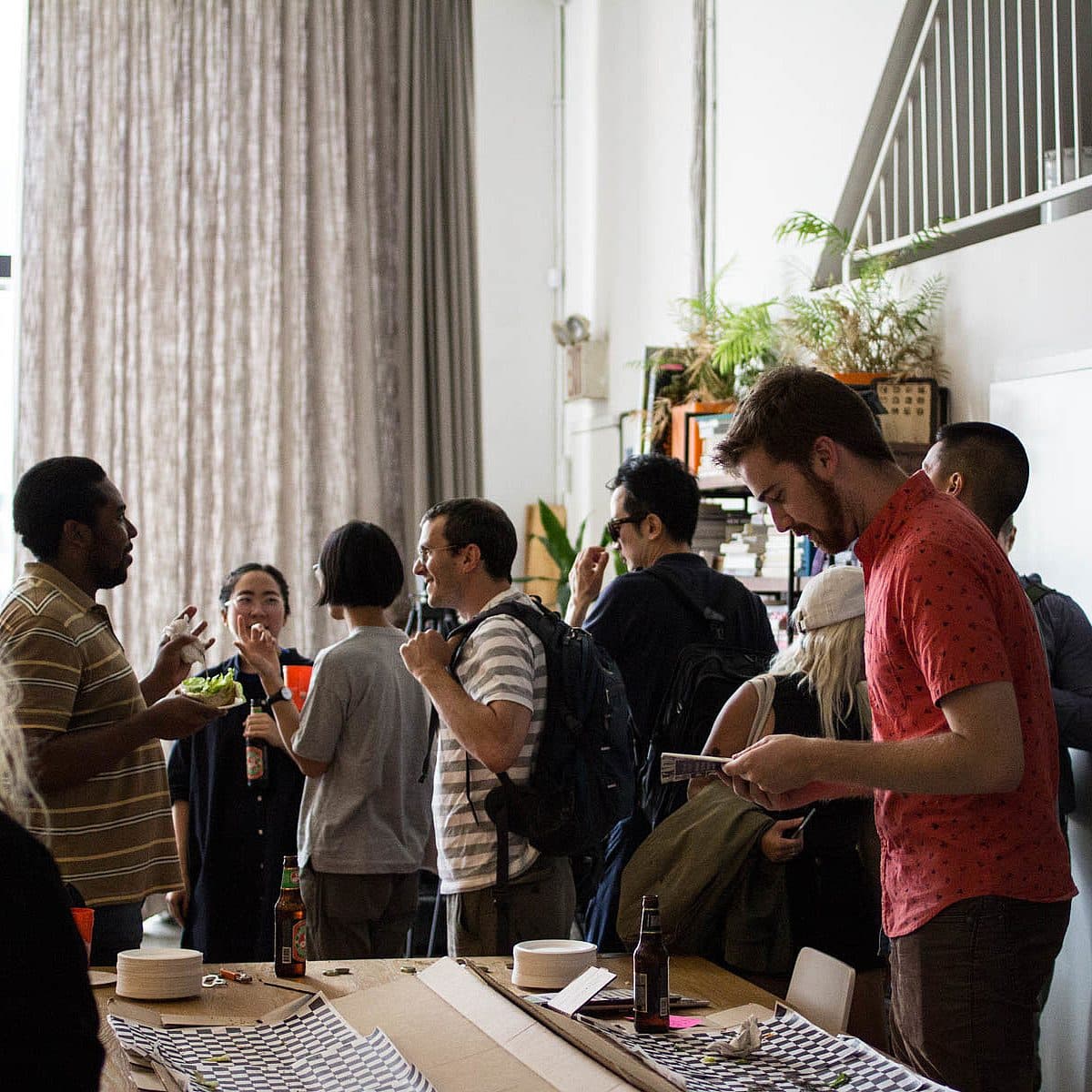 A group of people socialize and engage in a lively conversation in an indoor setting. The room has large windows and a high ceiling with a staircase. One person is writing in a notebook, while others are chatting and enjoying food. An indoor plant is visible in the background.