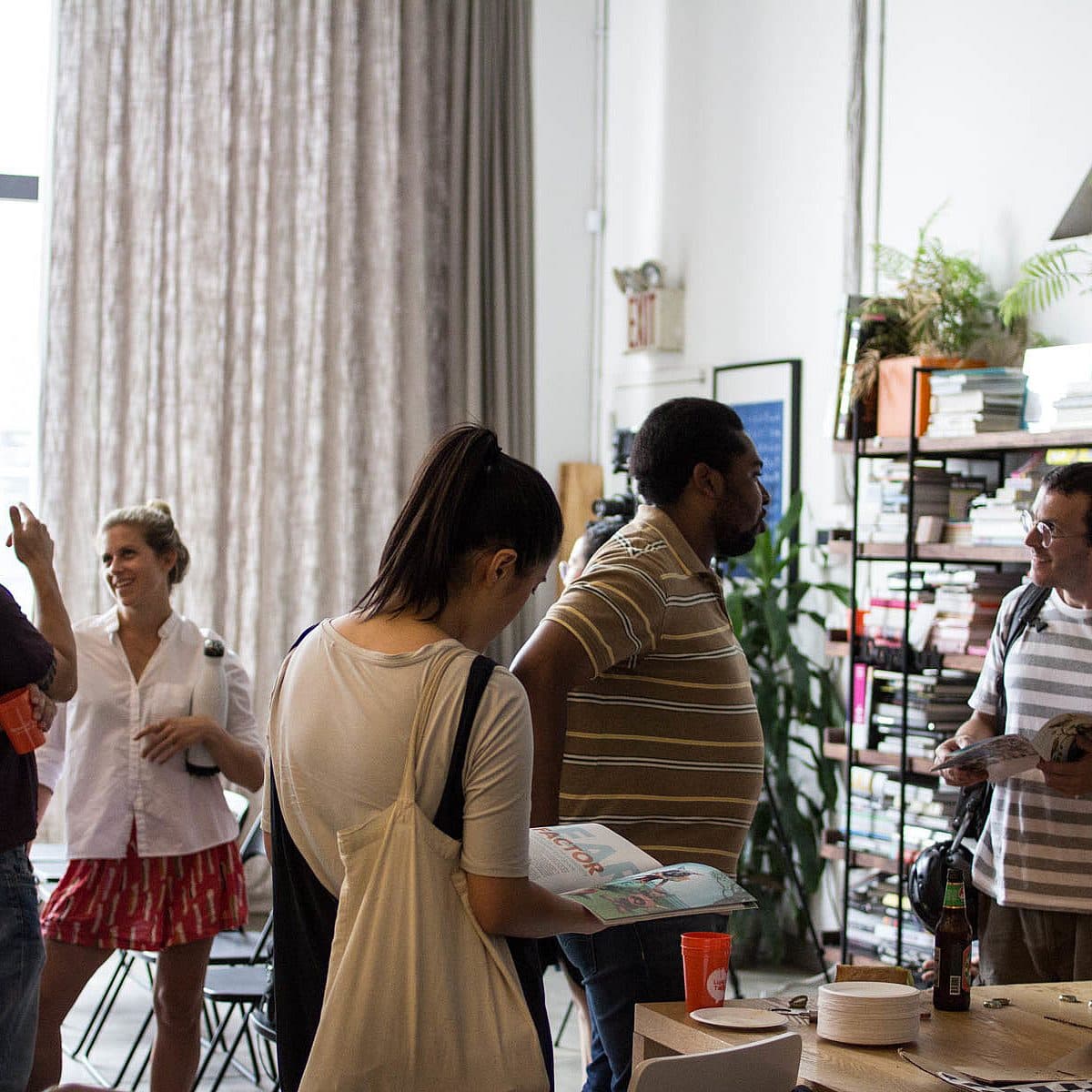 A group of people socializing in a well-lit room with a large window. Several individuals are holding drinks or brochures and are engaged in conversations. There's a bookshelf with numerous items and potted plants in the background.