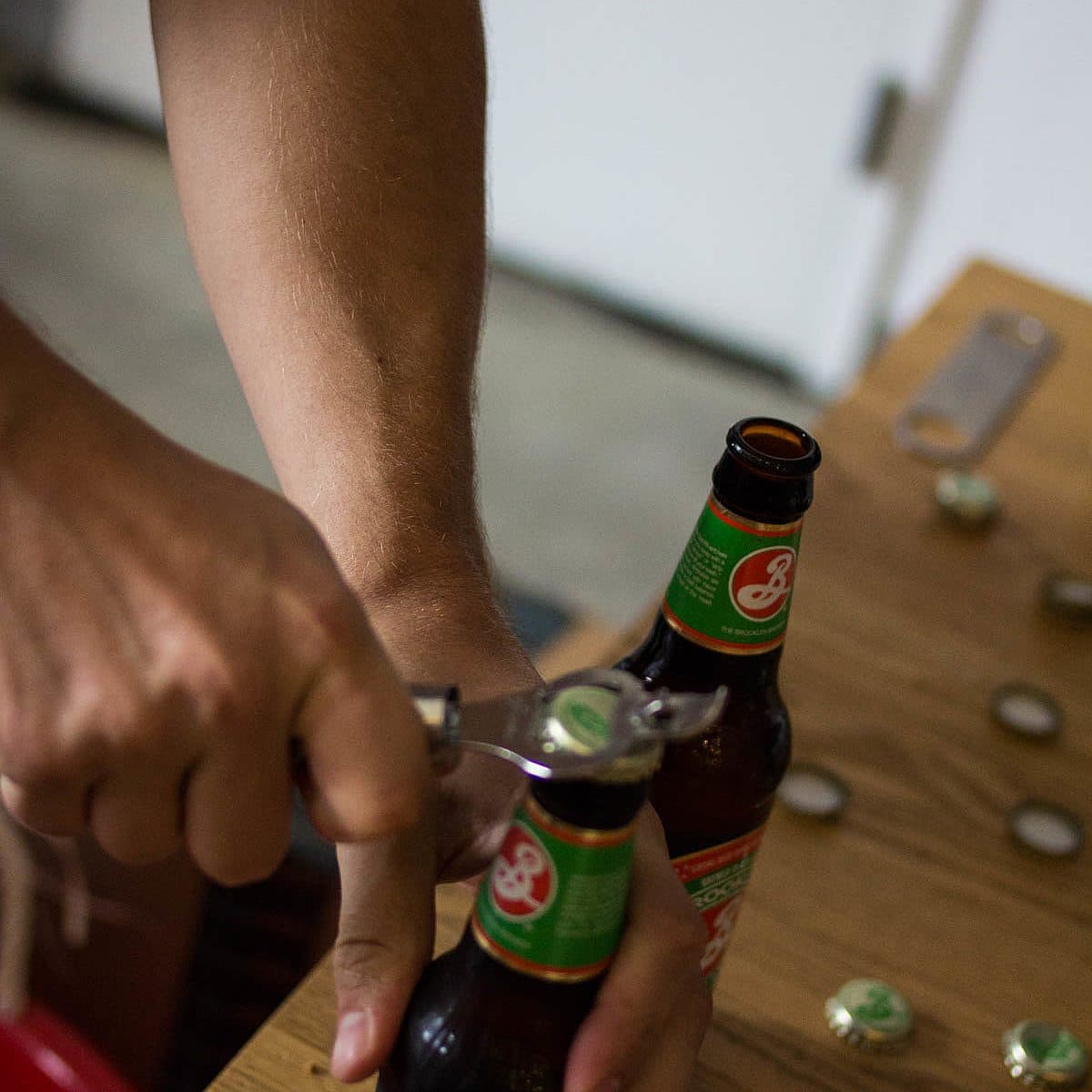 A person opening a bottle of beer with a bottle opener. The table in front of them has several bottle caps and another unopened beer bottle with a green and red label.