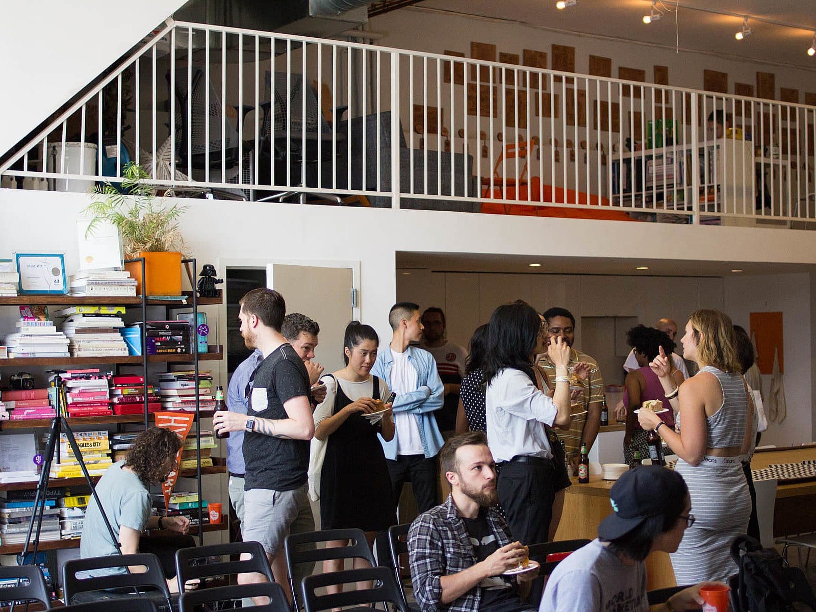 A group of people are gathered in an indoor space with modern decor, chatting and eating. A bookshelf filled with colorful books is on the left, and there is an upper level with a white railing. Empty chairs are in the foreground, and various items are scattered around.