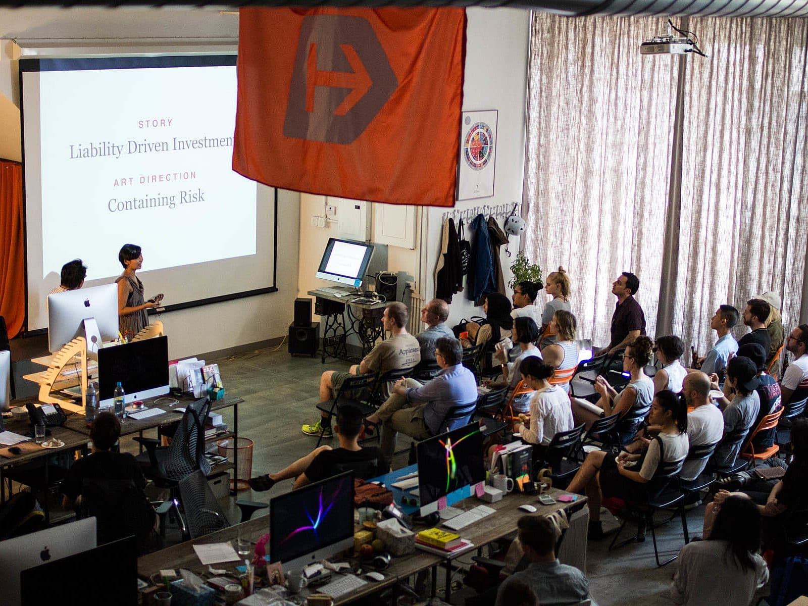 A group of people is seated in a room watching a presentation titled "Liability Driven Investments - Containing Risk." The room has computer screens, a large flag with an "H" on it, and various posters and items on the walls.