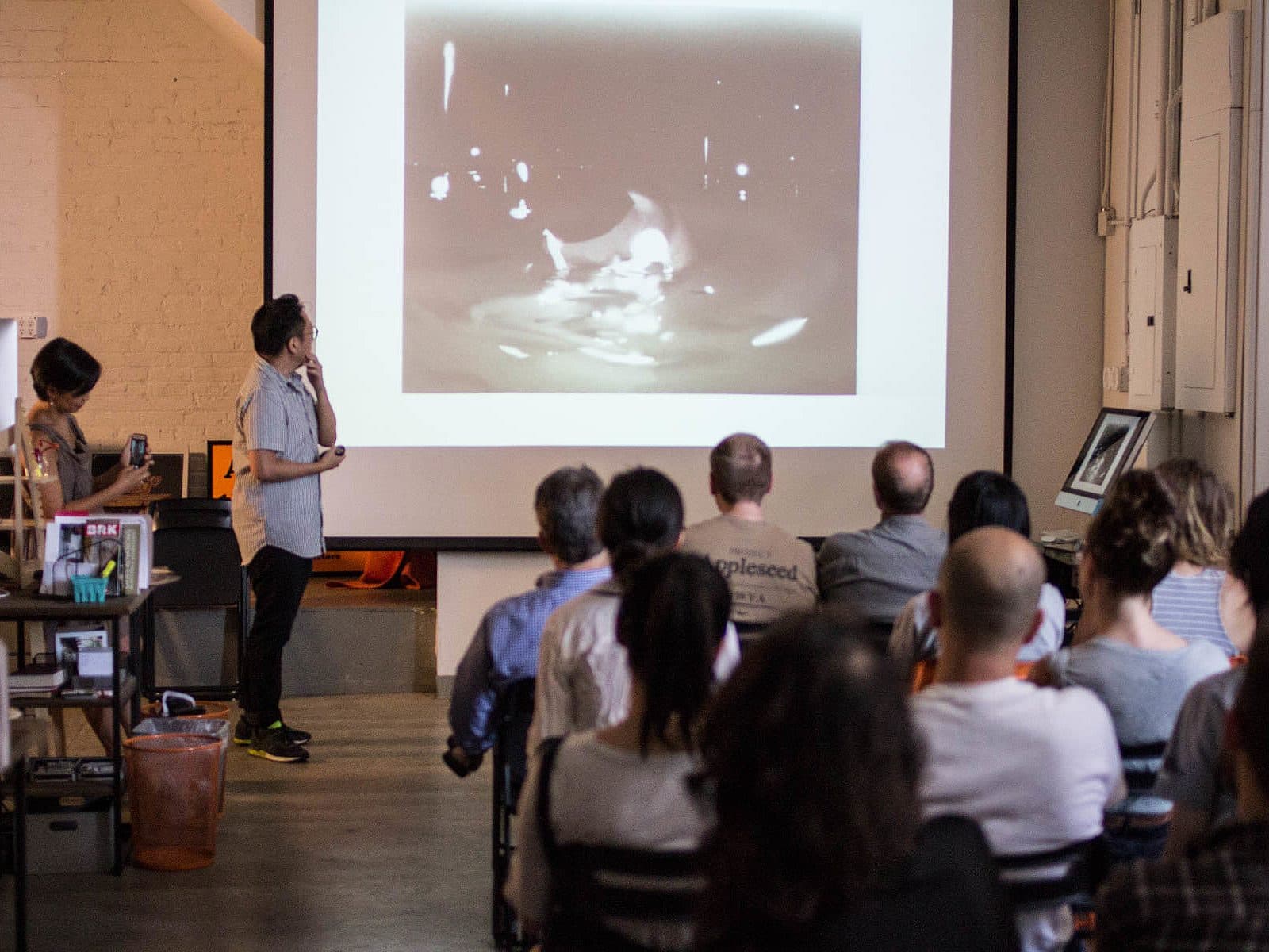 A person stands in front of a screen, presenting an image to an audience seated in rows. Another person nearby assists with the presentation. The room has a casual setting with a mix of art supplies and electronic equipment.