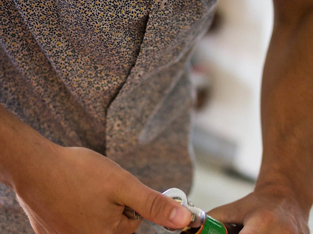 A person in a floral-patterned shirt is opening a bottle with a metal bottle opener. Only their torso and hands are visible as they twist the opener against the top of the bottle, which has a green and red label.