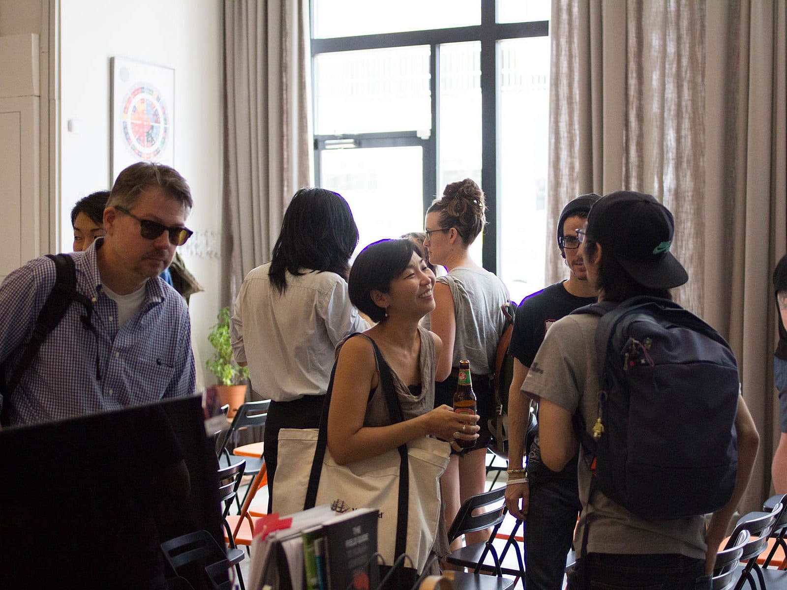 A group of people is casually chatting and mingling in a well-lit room with large windows and tall curtains. Some are holding drinks, and others have backpacks. One person is wearing sunglasses indoors, adding to the informal and relaxed atmosphere.