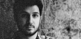 Black and white photo of a person with short dark hair and a beard, gazing directly at the camera. The background appears textured and slightly out of focus, giving a vintage or artistic feel to the image.