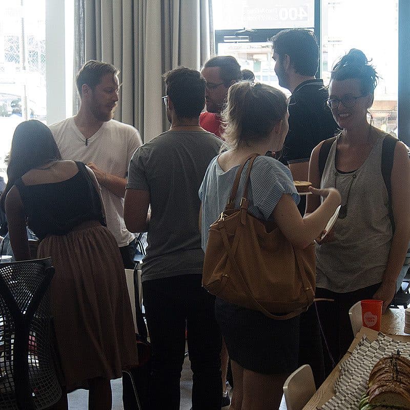 A group of people gathered in an office space, engaged in conversations. Some are standing near a table with food and drinks. Chairs and office equipment are visible, with large windows letting in natural light in the background. The atmosphere appears casual and social.