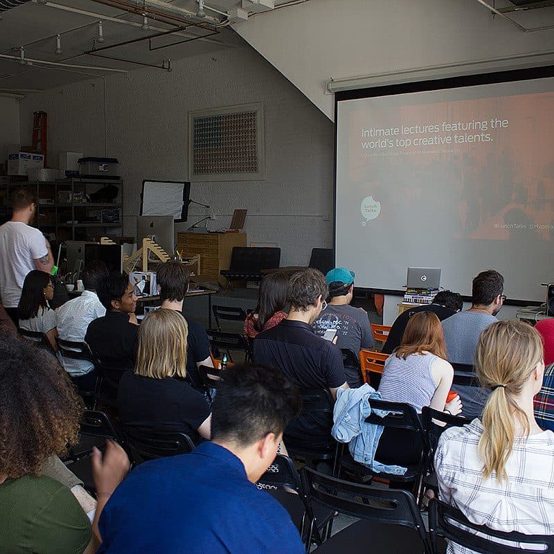 A group of people are seated in a classroom facing a screen displaying a presentation titled "Intimate lectures featuring the world's top creative talents." One person stands near the front, adjusting equipment. The room has a casual, industrial feel with various supplies and equipment in the background.