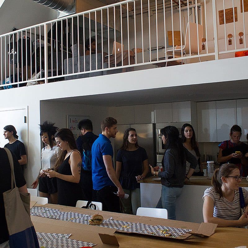 A group of people socializing in a modern kitchen area with a mezzanine level above. Some are standing and chatting, while others are seated at a long table with assorted food and drinks. The space has a mix of natural light and overhead lighting.