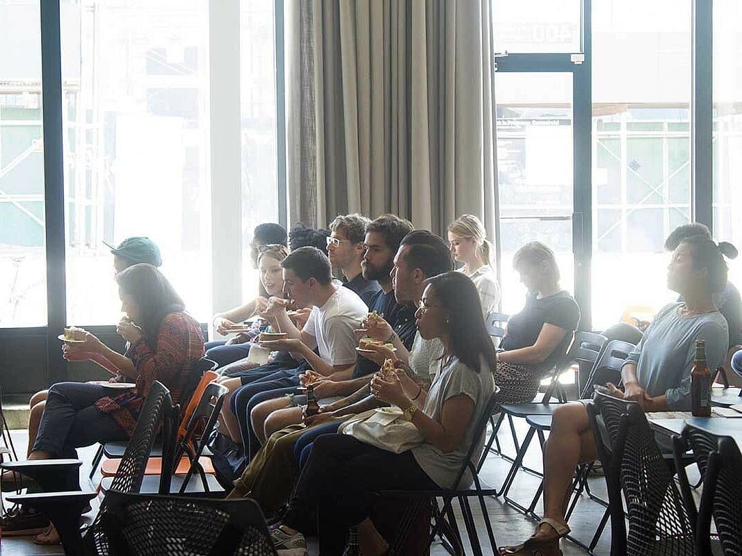 A diverse group of people sits in rows of chairs, attentively facing the front in a well-lit room with large windows. Some individuals are eating and drinking, while others appear to be listening intently. The atmosphere seems informal and relaxed.