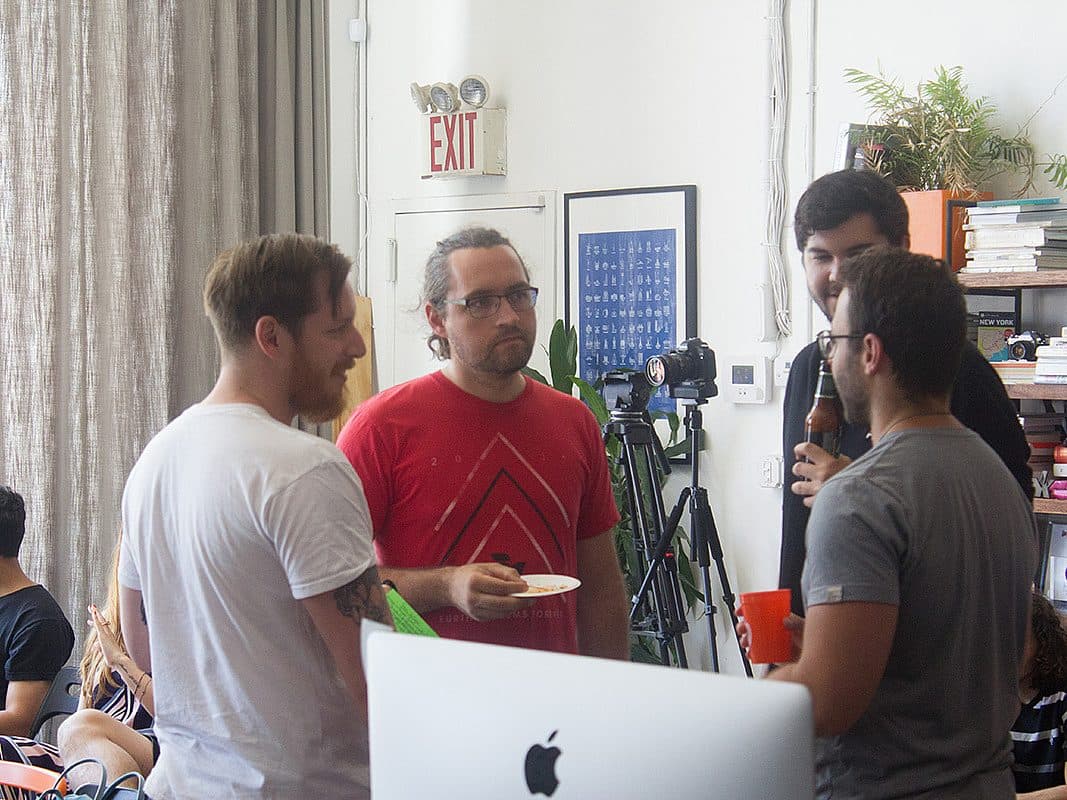 Four men are engaged in conversation in a casual indoor setting. One man in a red shirt holds a plate, while another in a gray shirt holds a drink. In the background, there are shelves with books and plants, a camera on a tripod, and a visible exit sign.