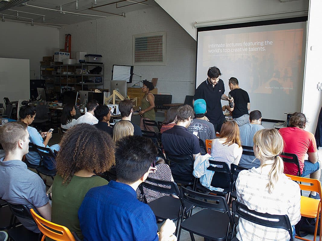 A group of people seated in rows facing a large screen displaying a presentation in a spacious room. Some individuals are standing and talking near the front. The audience appears to be attentively watching or listening.