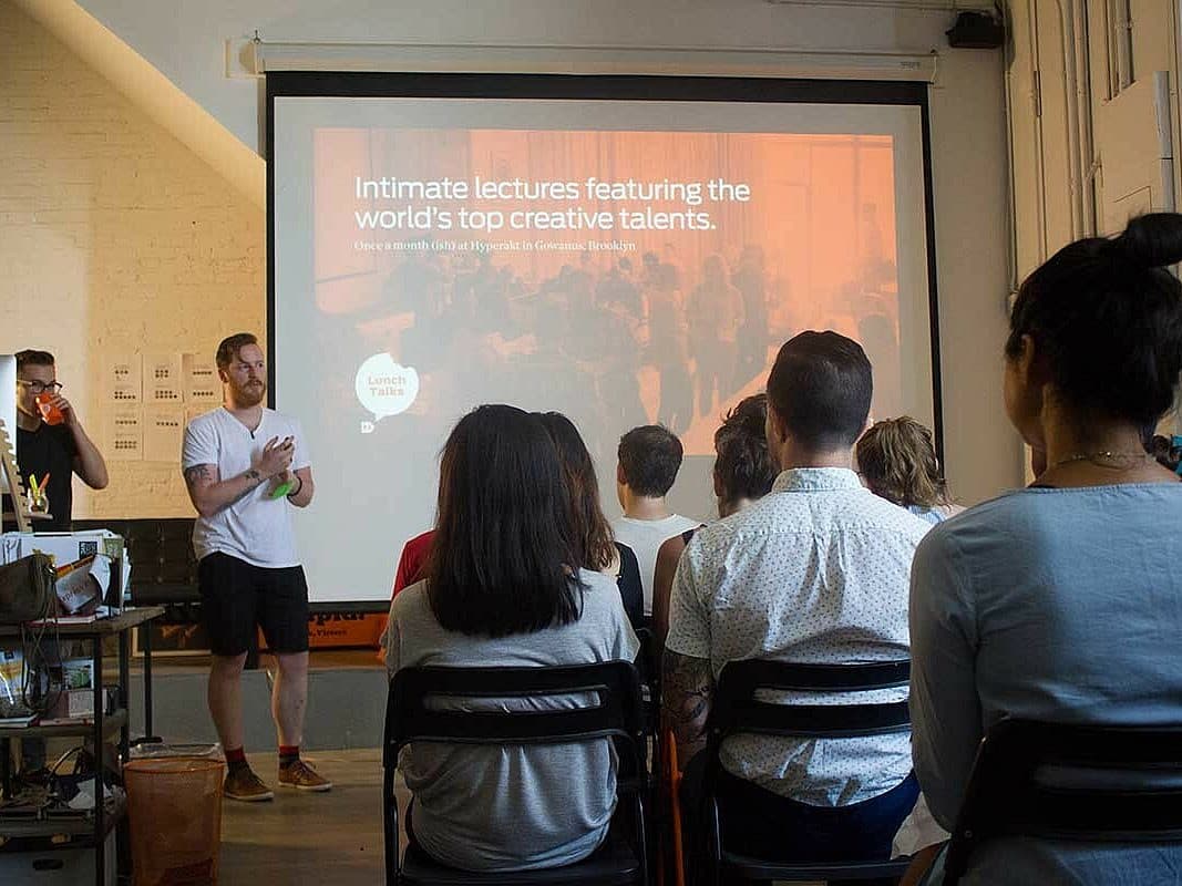 A small group of people sits in a room facing a large screen. A bearded man in a white shirt and black shorts stands to the left of the screen, speaking. The screen reads, "Intimate lectures featuring the world's top creative talents.