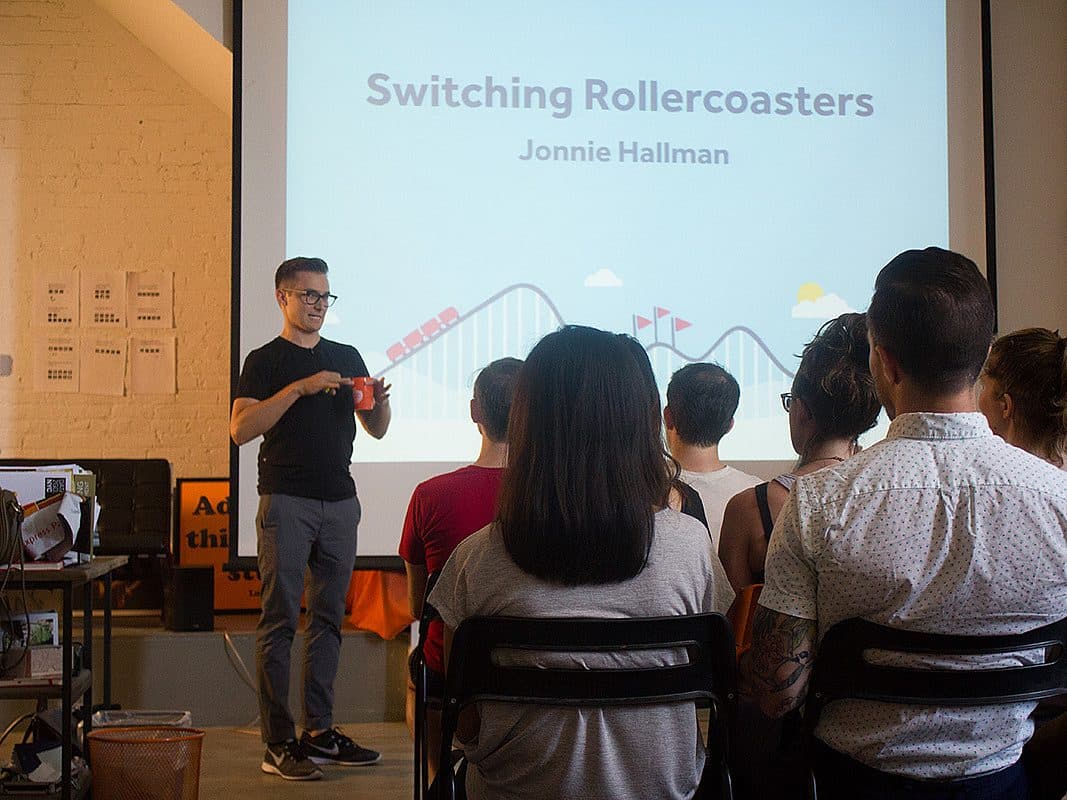 A person stands at the front of a room giving a presentation titled "Switching Rollercoasters" with a rollercoaster graphic on the screen behind them. Several people are seated, facing the presenter. The room has a bright ambiance with a projector screen and scattered items.