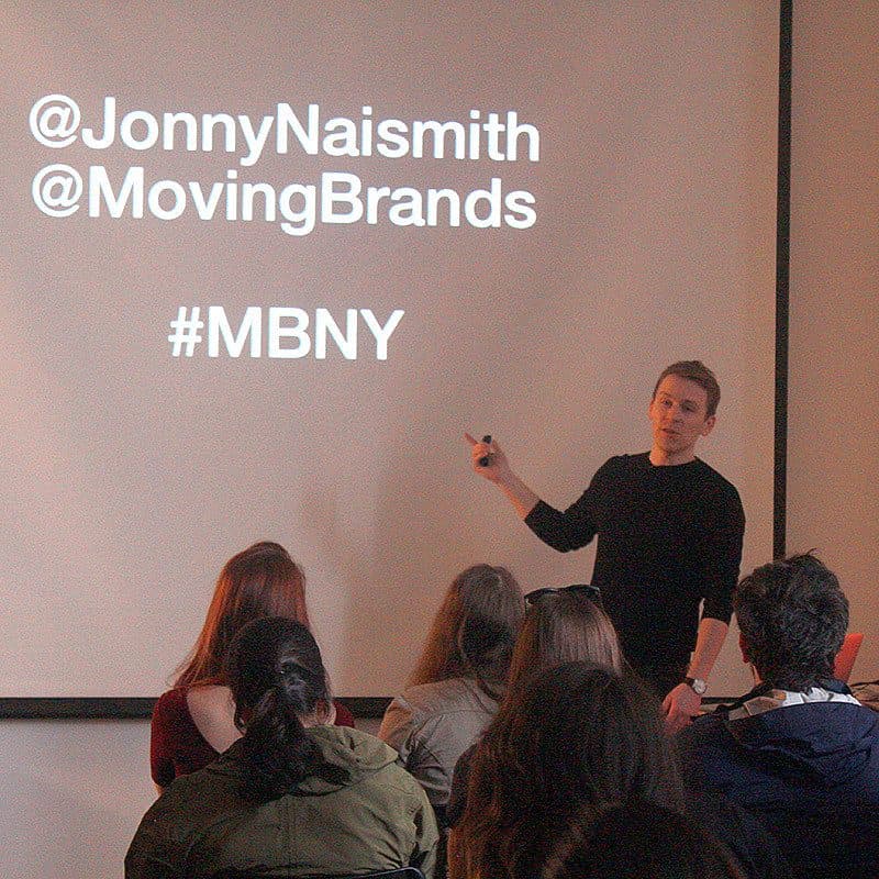 A person is standing in front of an audience, pointing at a screen. The screen displays the Twitter handles @JonnyNaismith and @MovingBrands, along with the hashtag #MBNY. The audience is seated, facing the speaker and the screen.