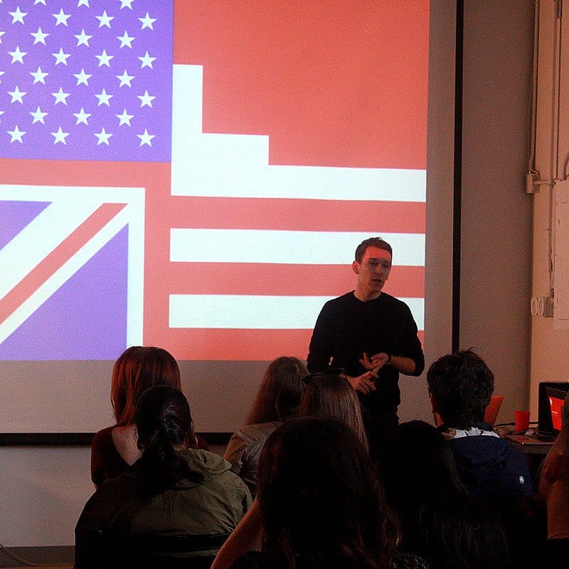 A speaker stands in front of a group of seated audience members. Behind the speaker is a large projection screen displaying a combined design of the UK and US flags. The room is dimly lit, and the speaker is gesturing with their hands while presenting.