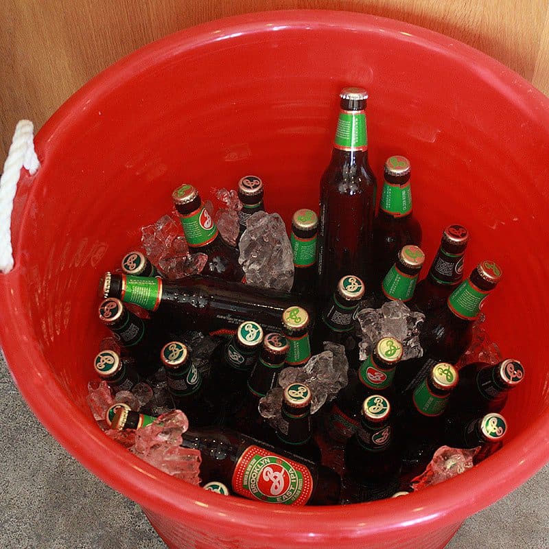A red bucket filled with ice and numerous green-bottled beers with red and gold labels. The bucket is placed on a concrete floor. Part of a person's lower leg wearing blue jeans and black Nike shoes is visible on the left side of the image.