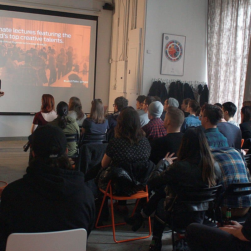 A speaker presents a lecture to a seated audience in a well-lit room. The projected slide reads, "Intimate lectures featuring the world’s top creative talents." The attendees face the presenter, who stands near the screen, engaging with the group.