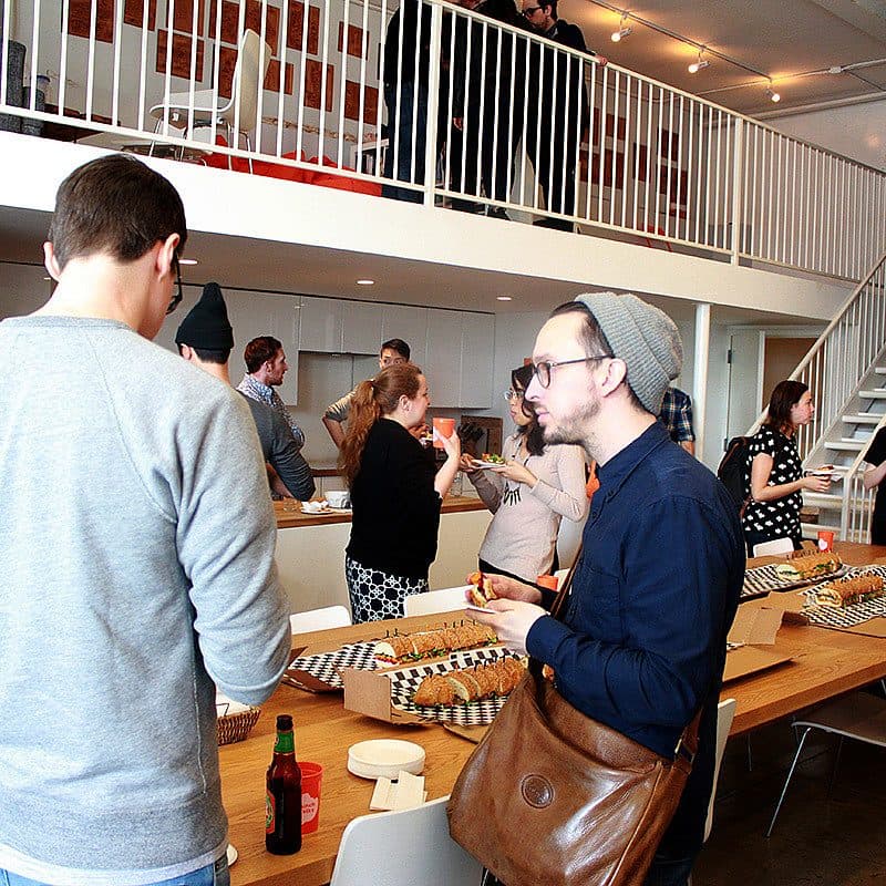 A group of people are gathered in a modern, open space with a mezzanine level. They are chatting and eating at a long table filled with food platters and pizza boxes. Some people are standing while others are seated. The atmosphere appears lively and casual.
