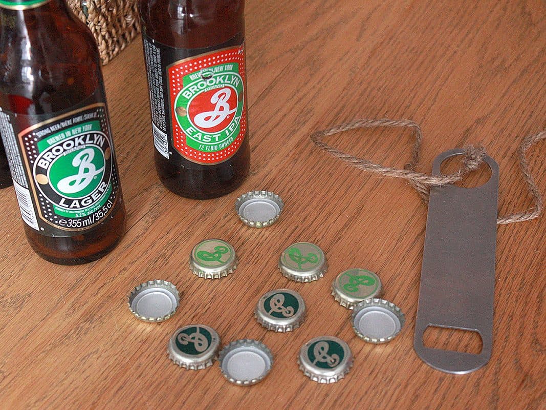 Close-up of two Brooklyn Brewery beer bottles, one with a green label marked "Brooklyn Lager" and one with a red label marked "Brooklyn East IPA," on a wooden surface. Several bottle caps, also branded with the brewery's logo, and a metal bottle opener with a twine loop are nearby.