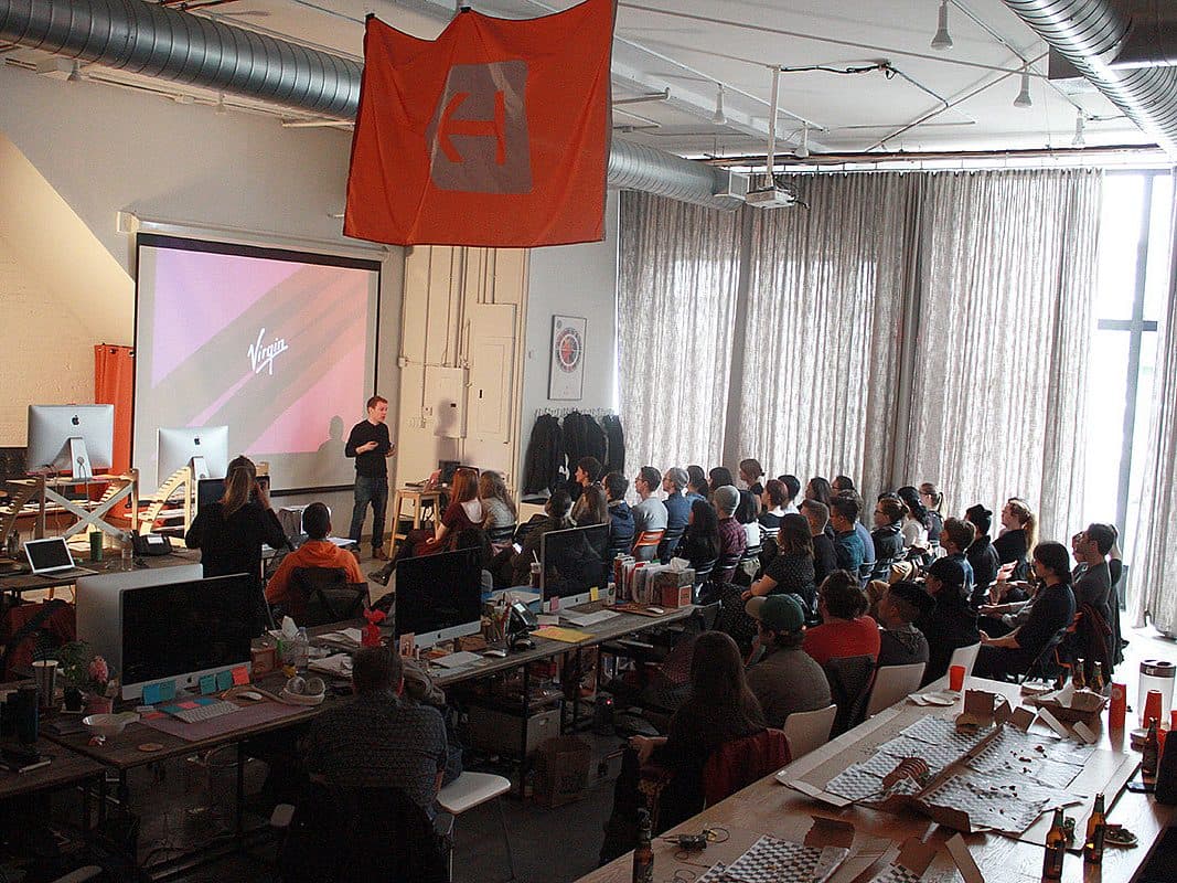 A group of people sit and listen to a presenter in an office space. The presenter is at the front near a large screen displaying a slide with text. Desks with computers and various items are in the foreground. A large orange flag hangs from the ceiling.