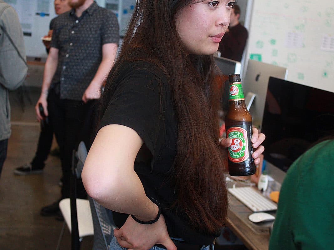 A woman with long dark hair stands at a desk holding a bottle of beer. She is wearing a black shirt and has a thoughtful expression. In the background, other people are engaged in conversation in what appears to be an office setting.