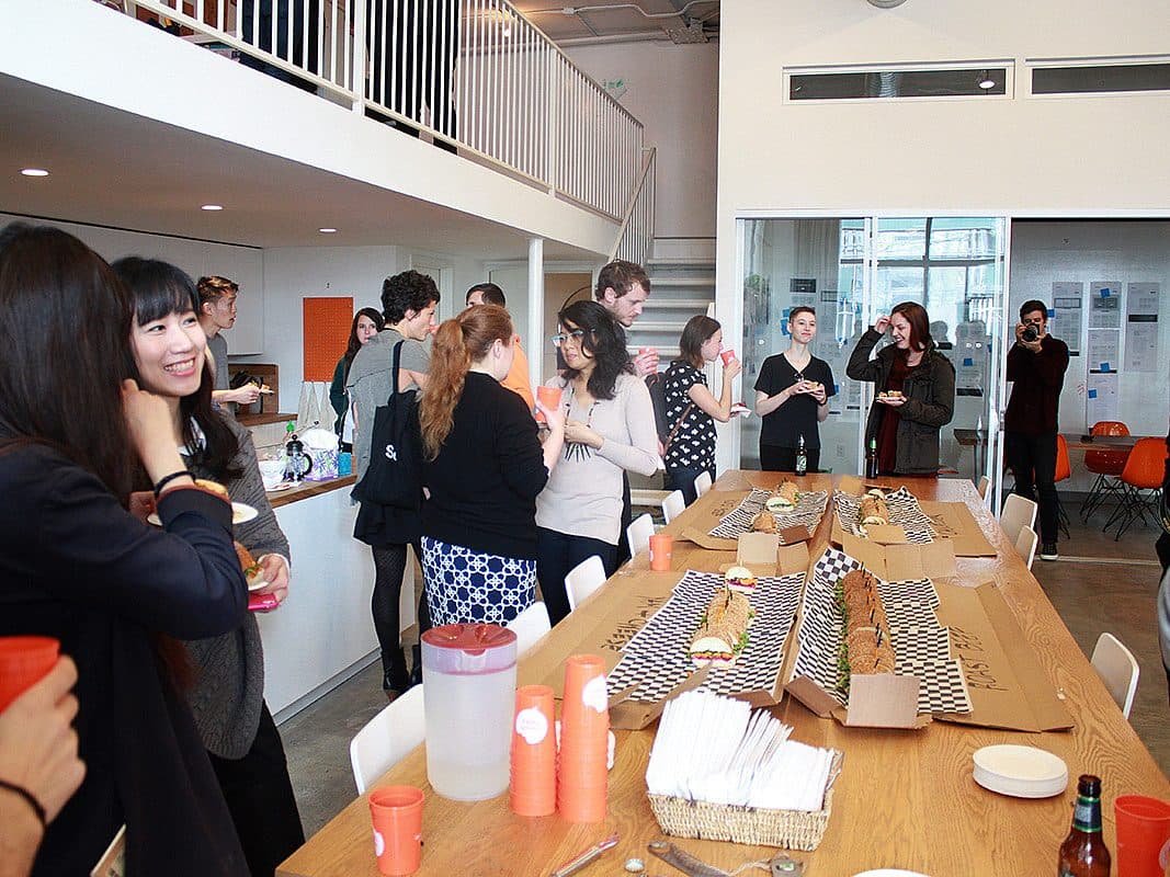 A group of people is gathered in a modern kitchen and dining space, engaging in conversations and enjoying a meal. A long wooden table is set with sandwiches, drinks, and disposable cutlery. The atmosphere appears lively and social.