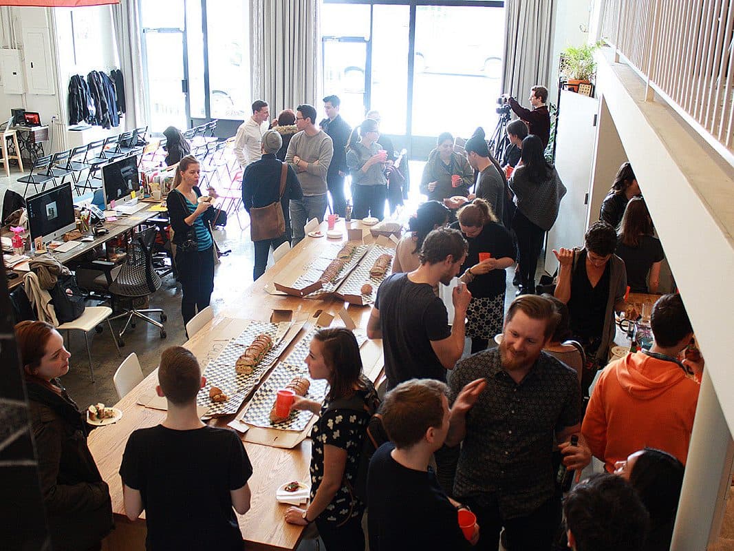 A large group of people gathered in an open office space, chatting and socializing. Long tables with assorted snacks and beverages are set up, and attendees are standing, eating, and engaging in conversations. Office equipment is visible in the background.