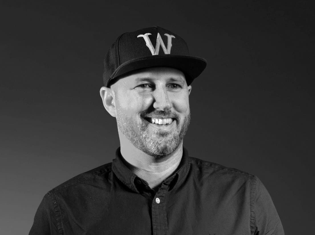 Black and white photo of a smiling person with a beard, wearing a baseball cap with the letter "W" on it and a button-down shirt. The background is plain and dark.