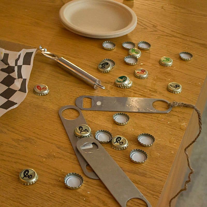 A wooden table with several bottle caps, a bottle opener, and a checked napkin. A small metal bottle opener and a disposable plate are also on the table. The bottle caps feature a black logo with a curly design on a gold background.