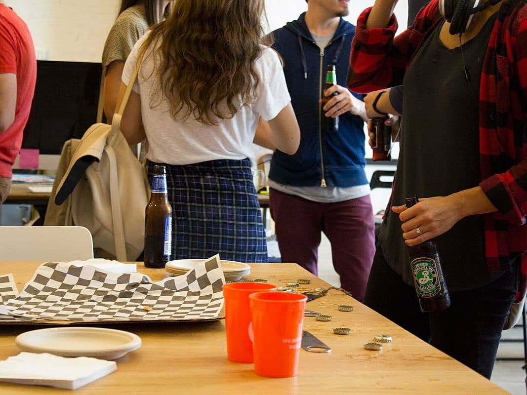 A group of people socializing at a casual indoor gathering. The focus is on a table with a checkered paper-lined tray, four orange cups, and bottle caps scattered around. Some individuals hold beer bottles, and one person is wearing headphones around their neck.