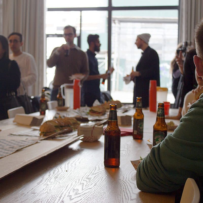 A group of people socialize in a room with large windows. In the foreground, a man in a green hoodie sits at a table with several beer bottles and food items. Others stand and chat in the background, creating a relaxed, casual atmosphere.