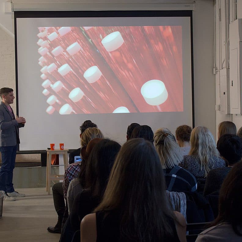A man stands in front of a seated audience giving a presentation. Behind him, a projection screen displays an image of red cylinders with white tops in rows. The room has white walls and some office and technical equipment visible.