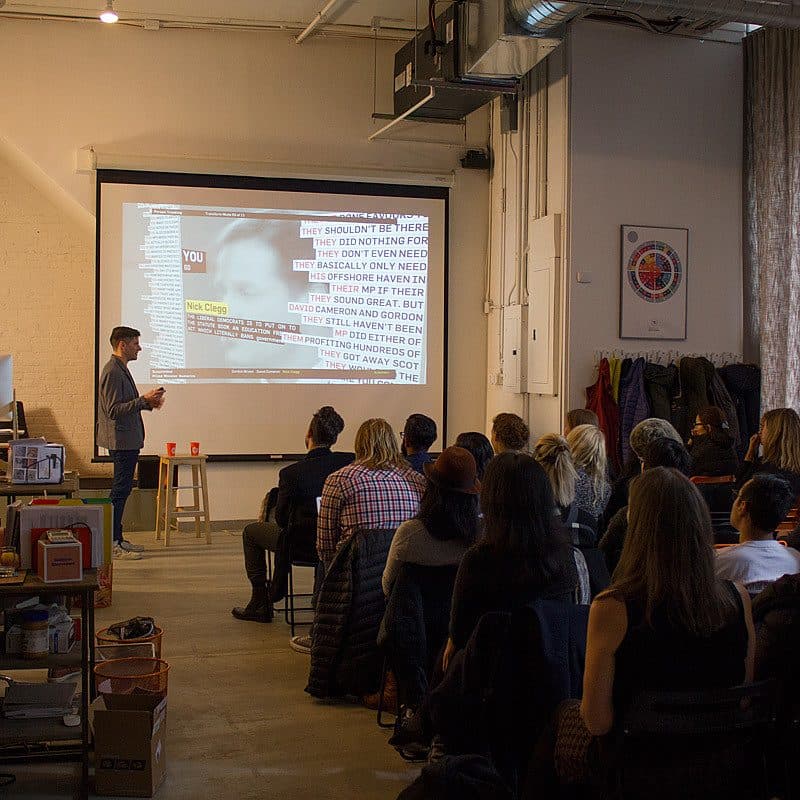 A person stands at the front of a room giving a presentation to a seated audience. The presentation includes a projected slide with text on a large screen. The room is well-lit with industrial decor, and attendees are attentively watching the presenter.