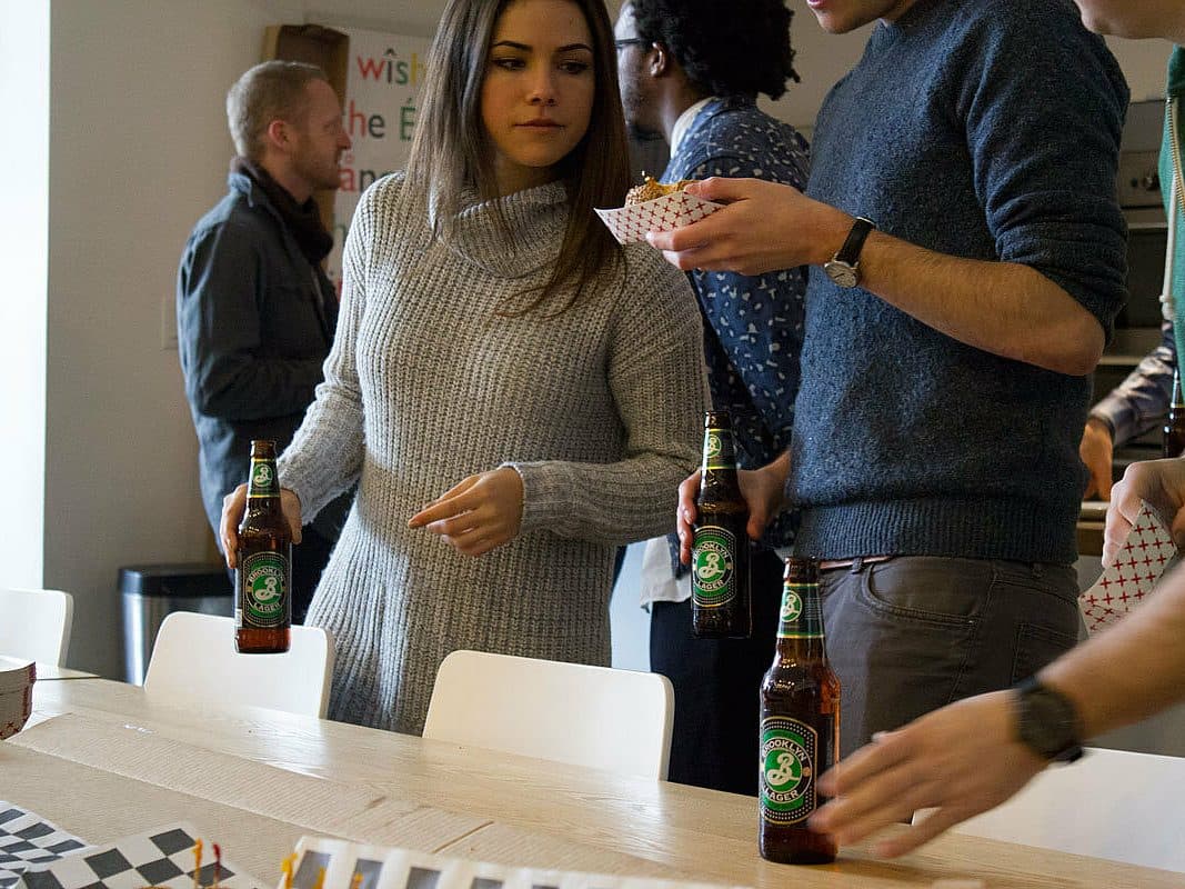 A woman in a gray sweater holds a beer bottle and looks to her right, while several people gather around a table holding food trays and drinks. The scene appears to be a casual gathering or party in an indoor space.