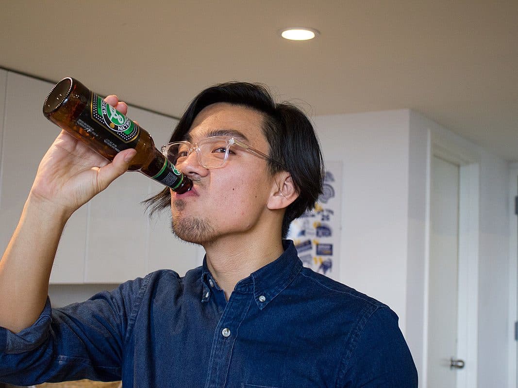 A person with glasses and a beard, wearing a dark denim shirt, is drinking from a bottle of beer. They are indoors in a modern kitchen with white cabinets and a partially open door in the background.