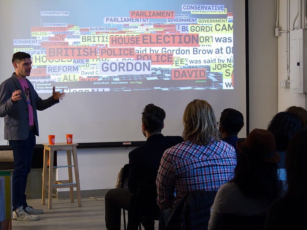 A speaker stands in front of an audience giving a presentation with a colorful word cloud projected on a screen. The word cloud features prominent words such as "BRITISH," "GORDON," and "ELECTION." The audience is seated, watching attentively.