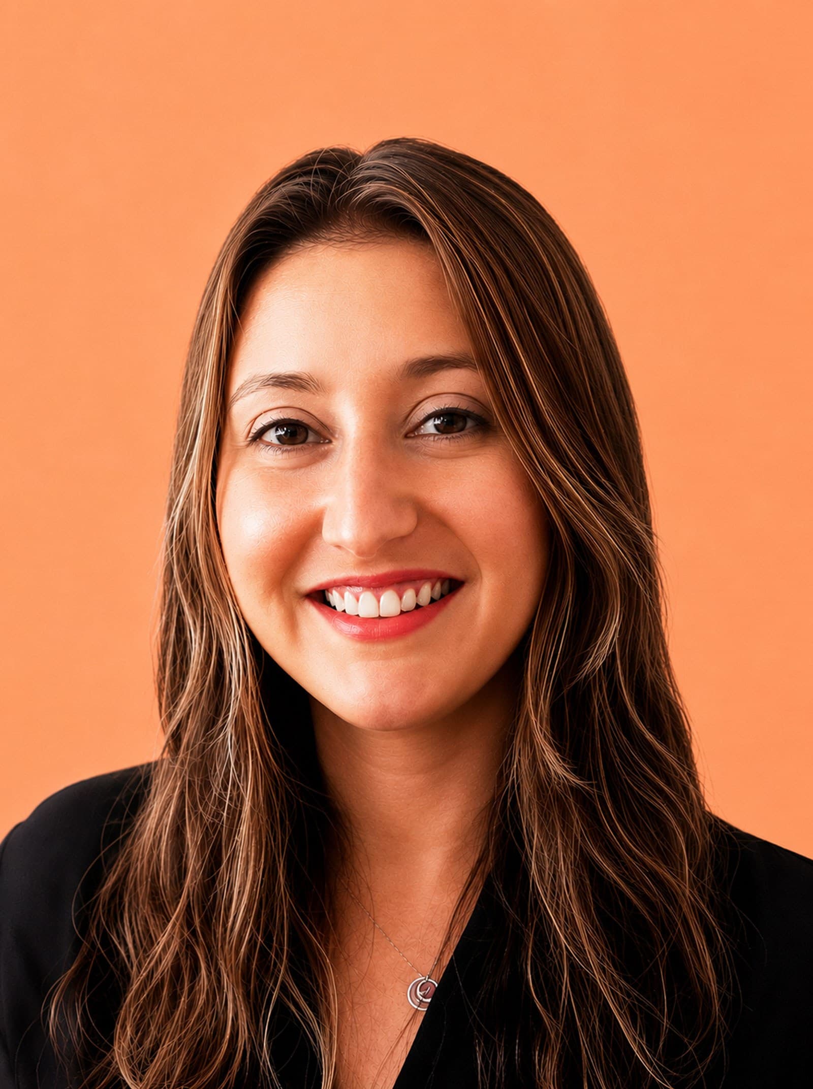 A woman with long brown hair smiles at the camera. She is wearing a black top and a necklace, with an orange background behind her.