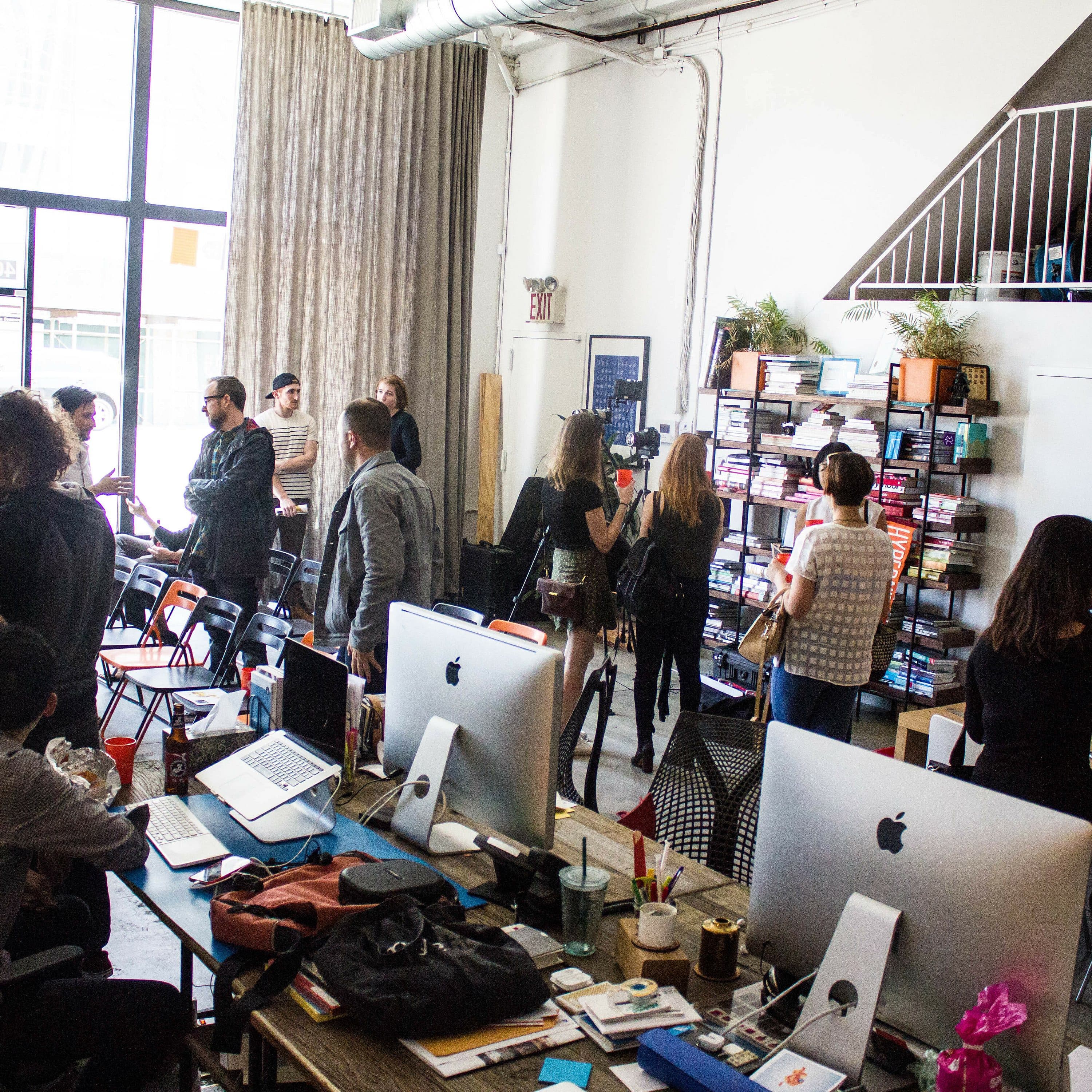 A modern office space with high ceilings and large windows. Several people are standing and talking in groups, while some are seated at desks with computers. Bookshelves are visible on the wall, and light streams in from the windows, creating a bright atmosphere.