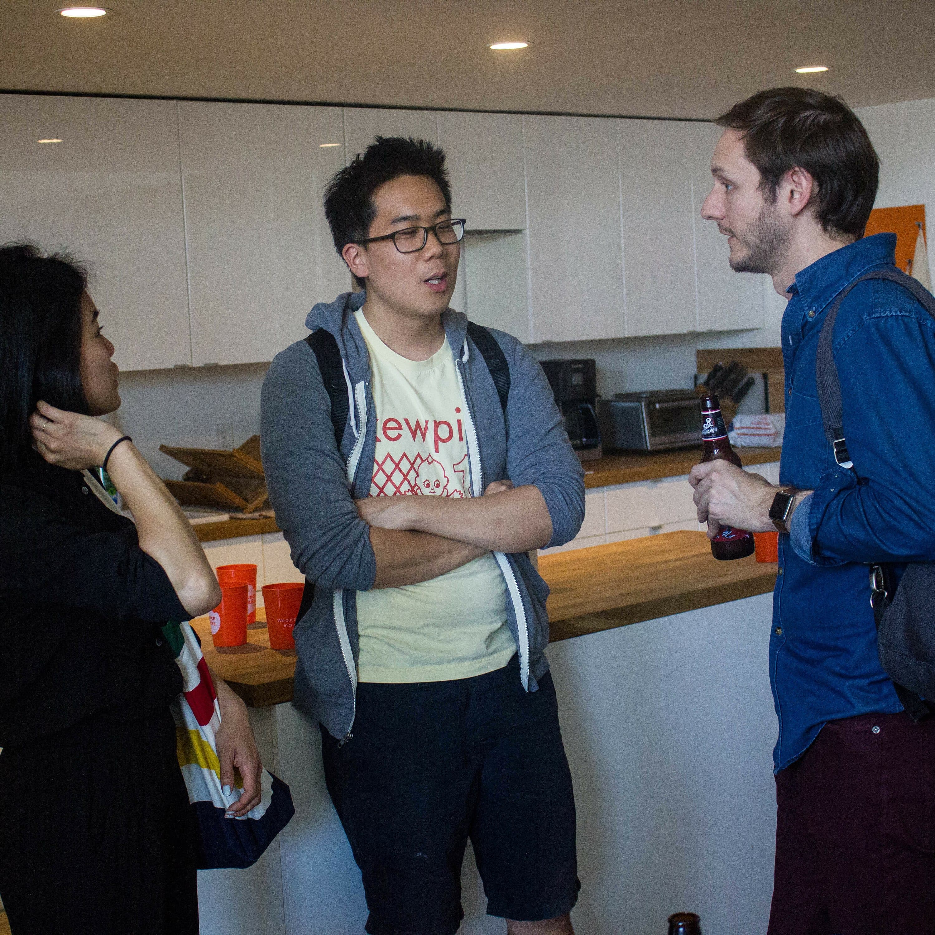 Three people are standing in a modern kitchen, engaged in conversation. One person is wearing a black outfit with a colorful bag, another is in a light shirt and gray hoodie, and the third is in a blue shirt with a backpack, holding a bottle.