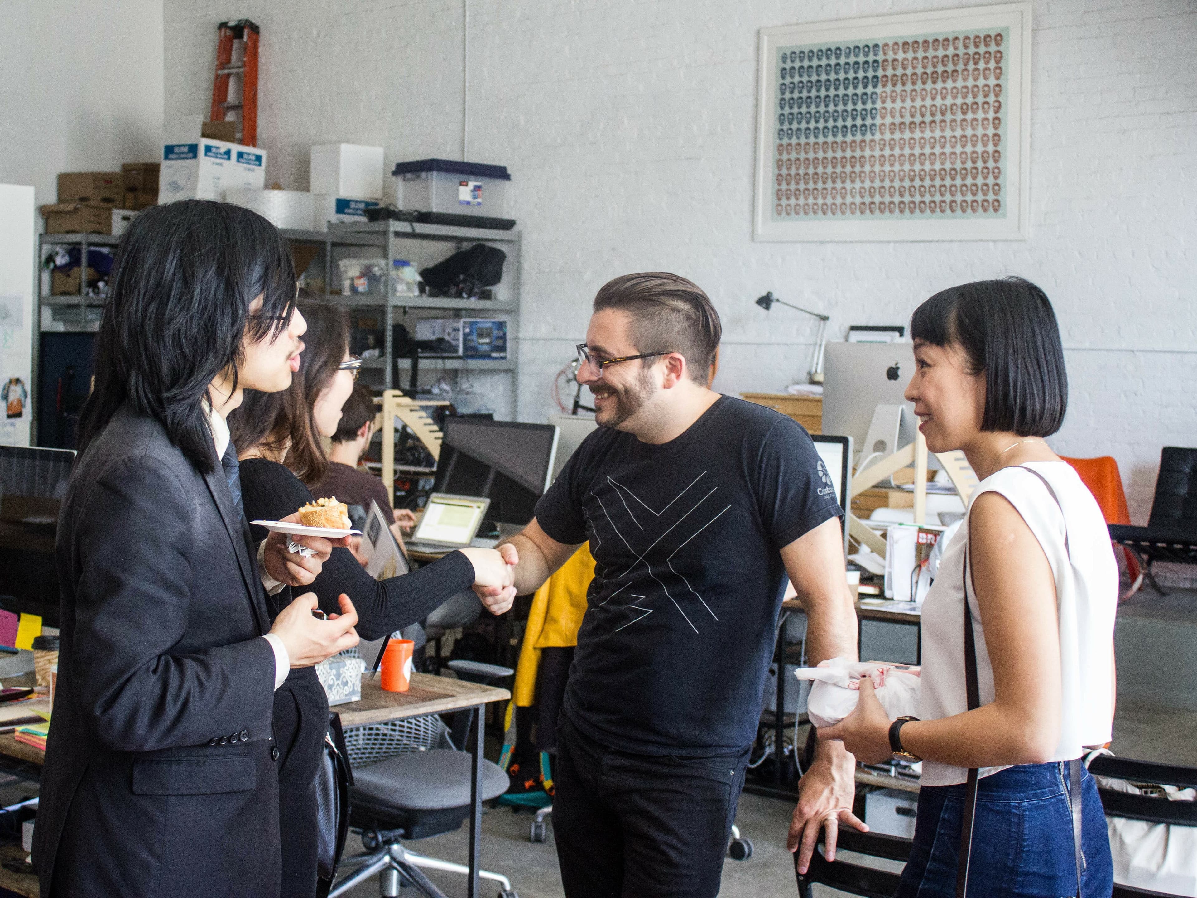 A group of three people is engaged in conversation at a casual office gathering. One person is holding a plate with food while another is shaking hands. The room is filled with desks, computers, and shelves with various items, creating a relaxed workspace atmosphere.