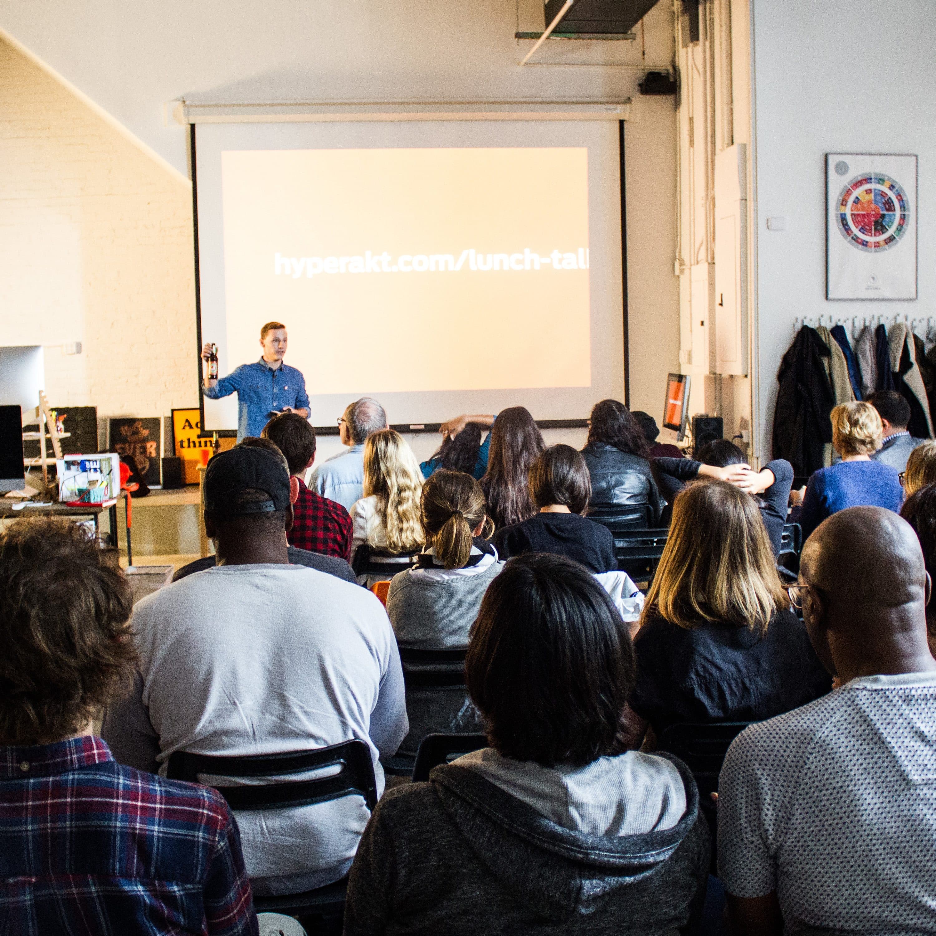 A group of people sit in chairs facing a projector screen displaying a URL (hyperakt.com/lunch-talk) in a modern, bright room. A person stands at the front giving a presentation, and various office items and artwork are visible around the space.