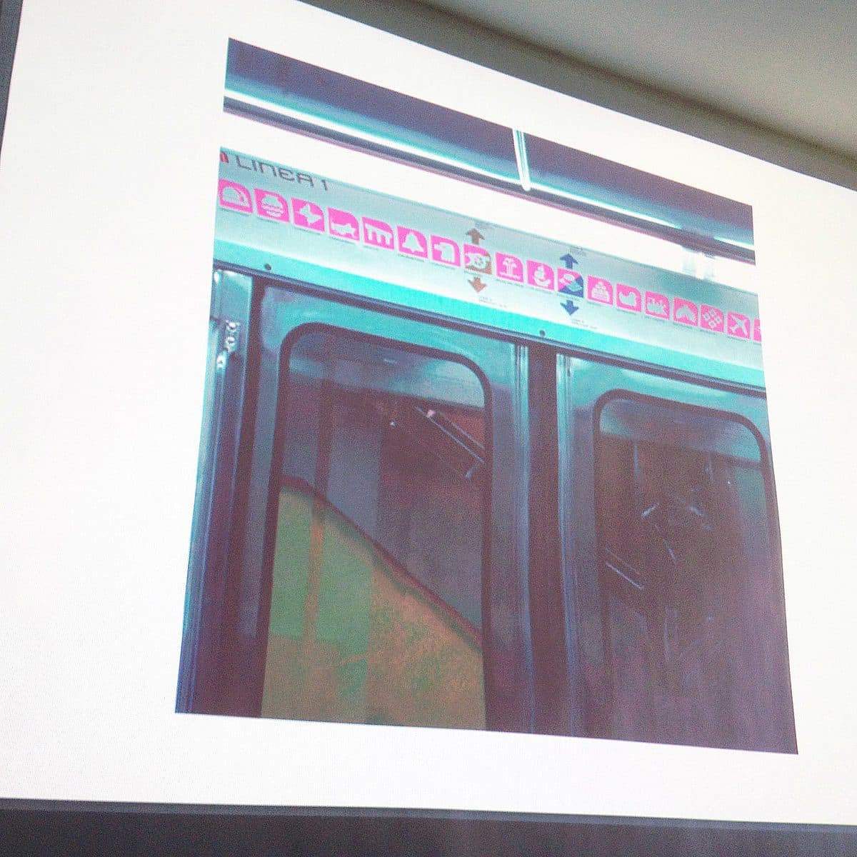 An image of a projected photograph depicting subway doors. Above the doors, there is a white strip with a series of pink symbols, possibly warning or informational signs. The projection is displayed on a white wall with slight shadows on the edges.