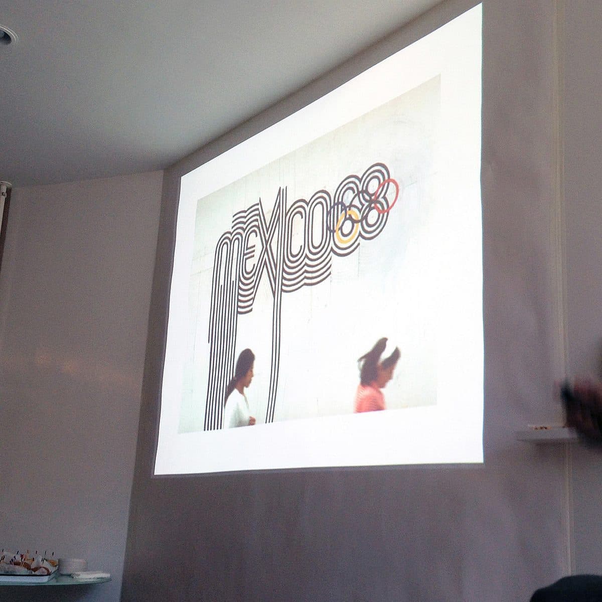 A person standing in a dimly lit room is pointing at a projector screen displaying artwork. The artwork features the word "My Love" styled in flowing, intertwined lines. A poster on the wall next to the projector screen reads, "I wish the English language had more diacritic characters.