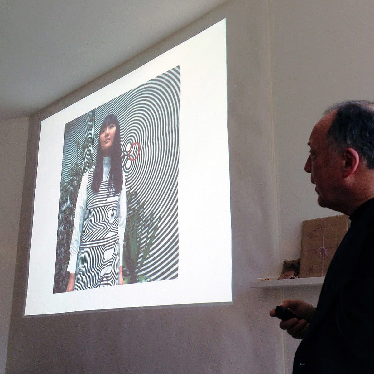 A person is giving a presentation, pointing to a projected image on a white screen. The image on the screen shows a woman standing in front of a black and white spiral pattern. The presenter appears to be wearing a dark jacket and is holding a pointer or remote.