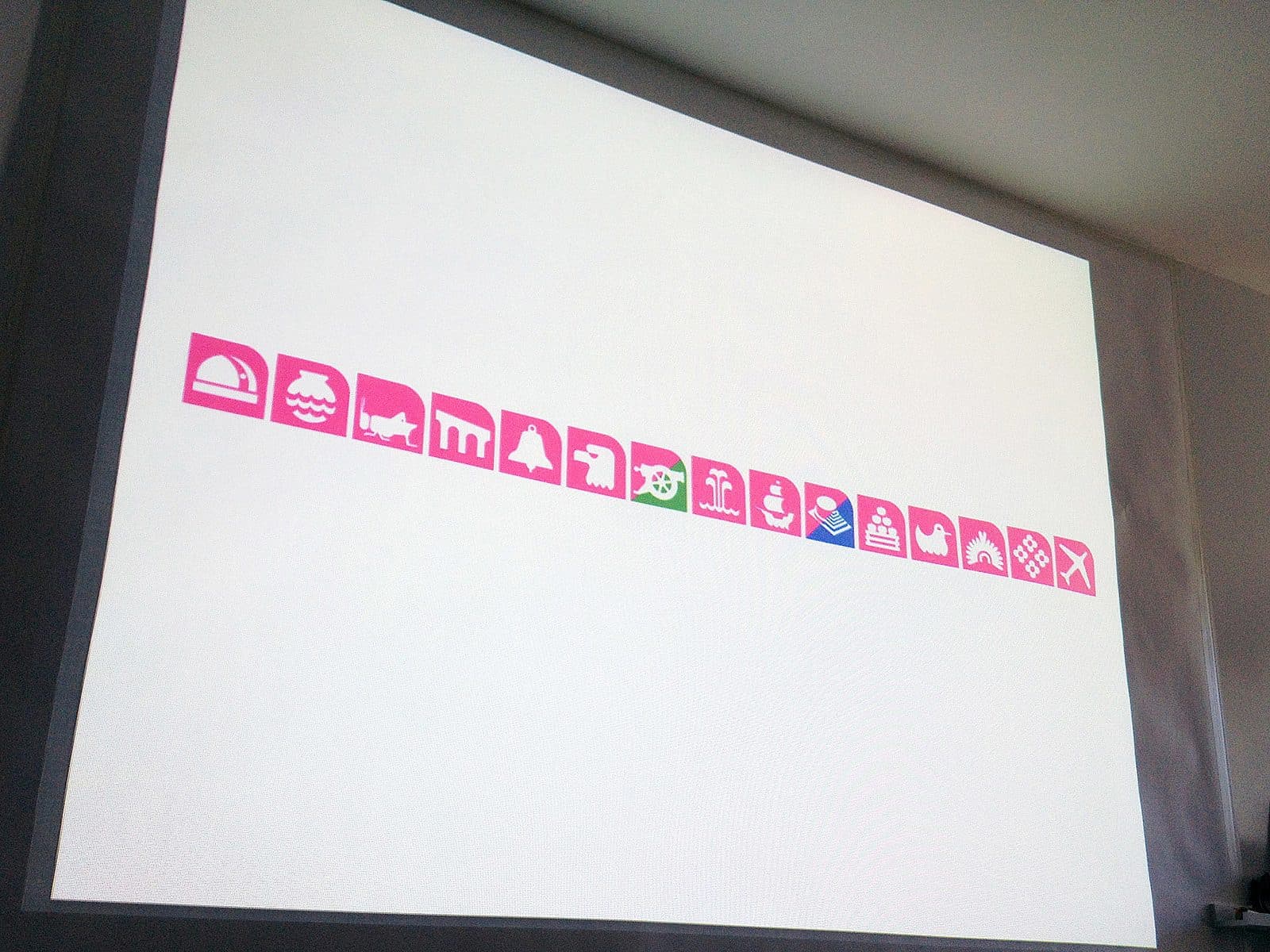 A horizontal row of pink icons is displayed on a white screen. Each icon contains a white symbol, including images of buildings, a mask, a cutlery set, a family with children, and various other symbols. The background is a neutral-colored wall.